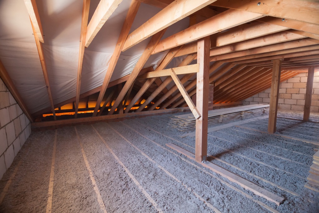 The image shows an unfinished attic space with exposed wooden roof beams and rafters, insulation material on the floor, and brick walls visible on one side.