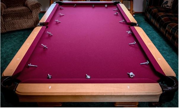 The image shows a burgundy-colored pool table with wooden rails and several white cue balls scattered on the felt surface, situated in what appears to be a basement or recreation room with carpeting and furniture visible in the background.