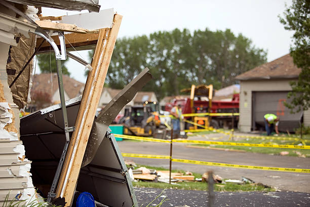 The image shows the aftermath of what appears to be structural damage to a building, with debris and wooden beams scattered around, yellow caution tape cordoning off the area, and emergency vehicles visible in the background.