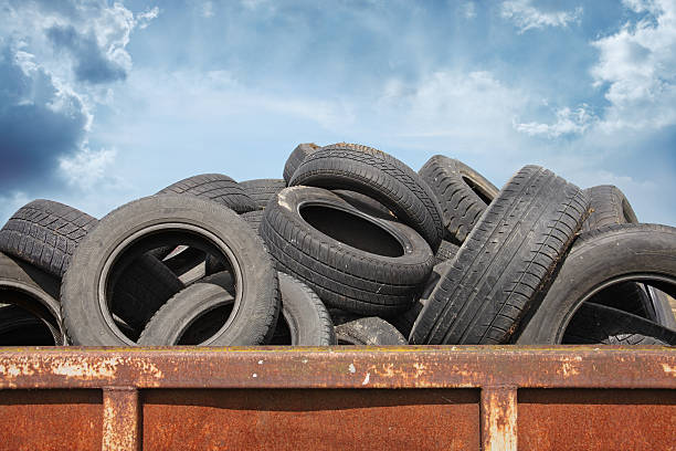 The image shows a large pile of old, used tires stacked in what appears to be a rusty metal container or dumpster against a cloudy blue sky.