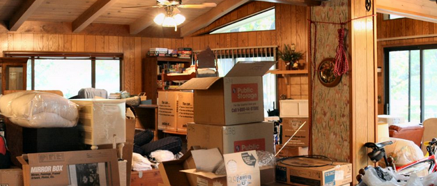 The image shows a cluttered interior room with wood-paneled walls and exposed ceiling beams, filled with numerous cardboard moving boxes and packed items, suggesting someone is in the process of moving or storing belongings.